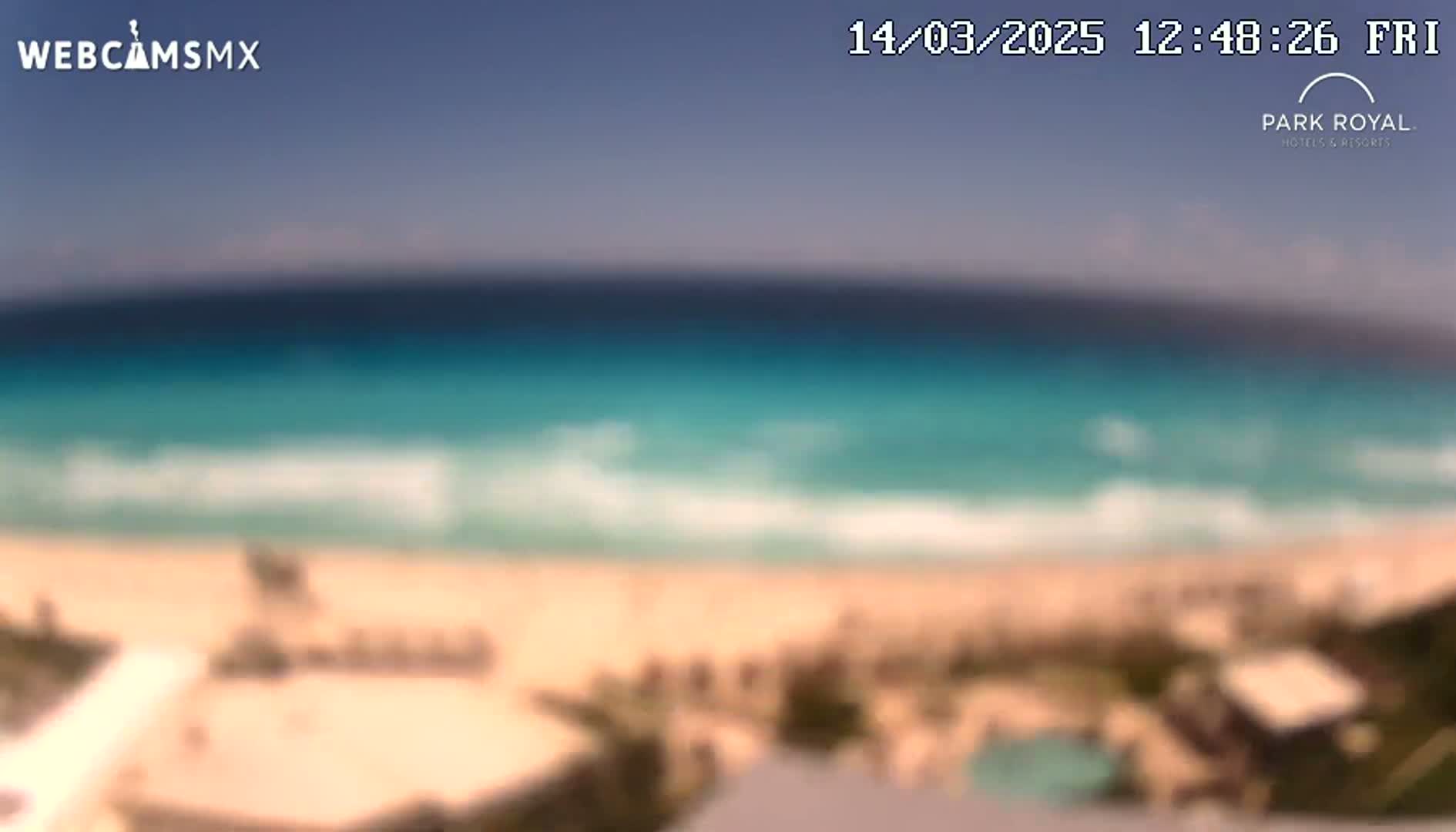 A blurry, high-angle view shows a beach with turquoise water and small waves under a clear sky.