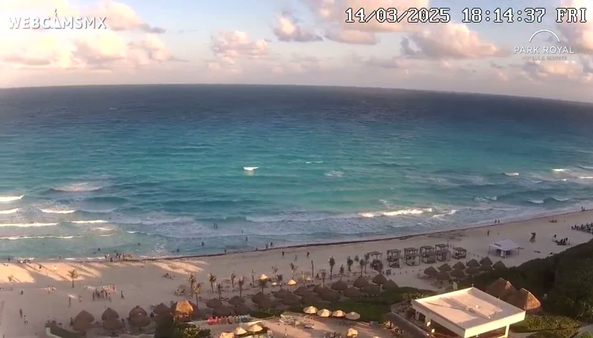 An expansive, turquoise ocean with small waves laps a sandy beach lined with beach umbrellas and a resort under a partly cloudy sunset sky.