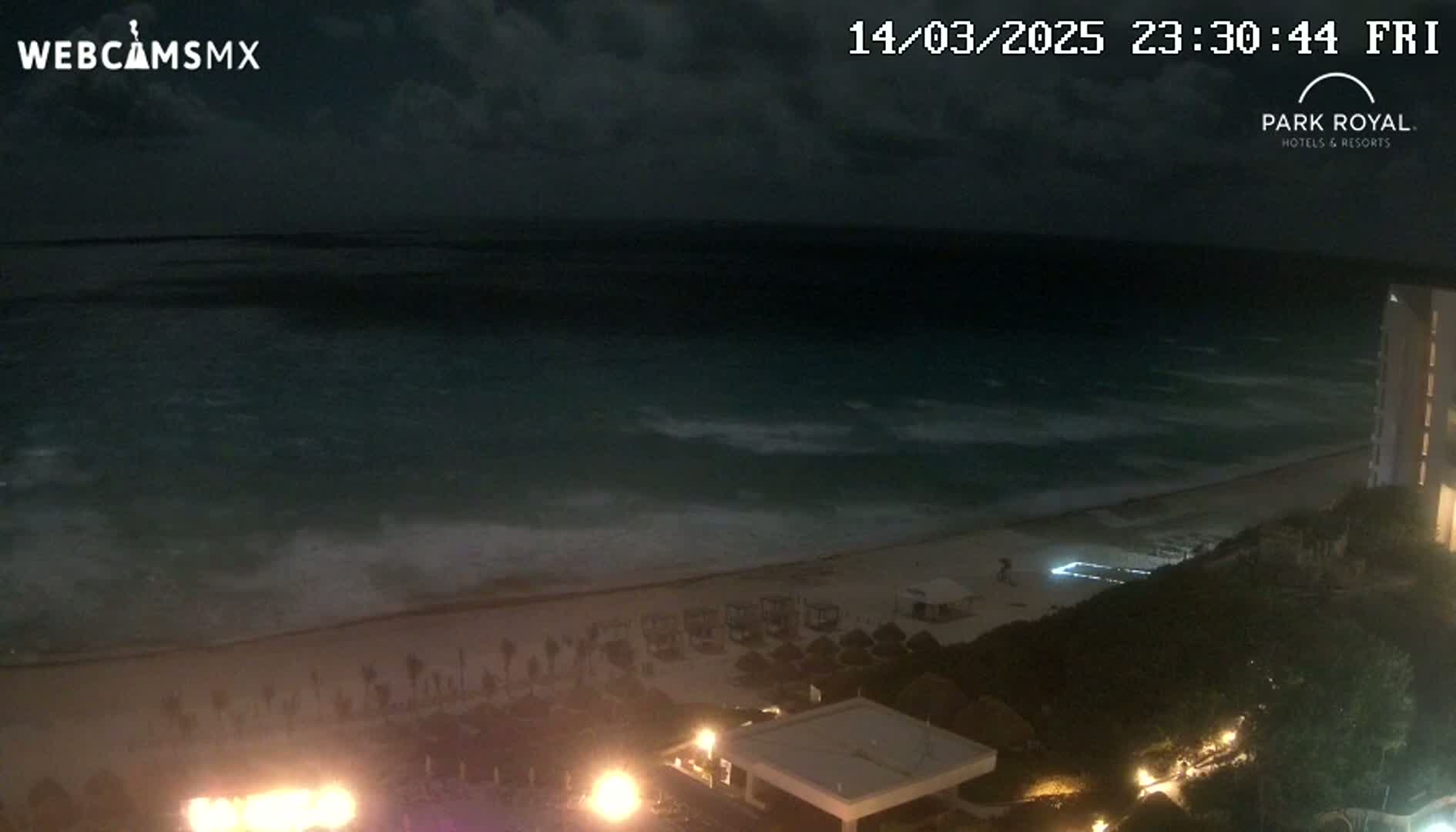 A nighttime beach scene shows a dark ocean with small waves lapping a sandy shore, where lights illuminate beach umbrellas and a resort building.