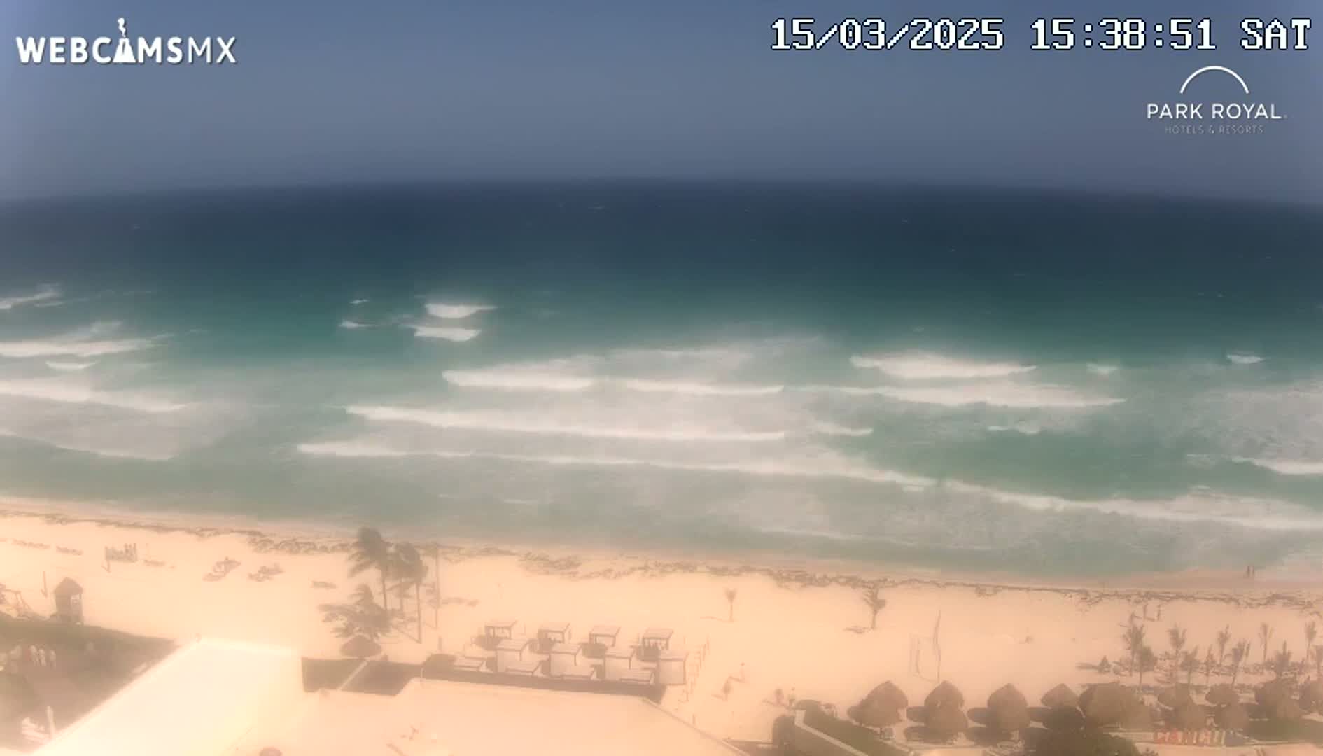 A sandy beach with small waves and a few palm trees is visible under a mostly clear sky.