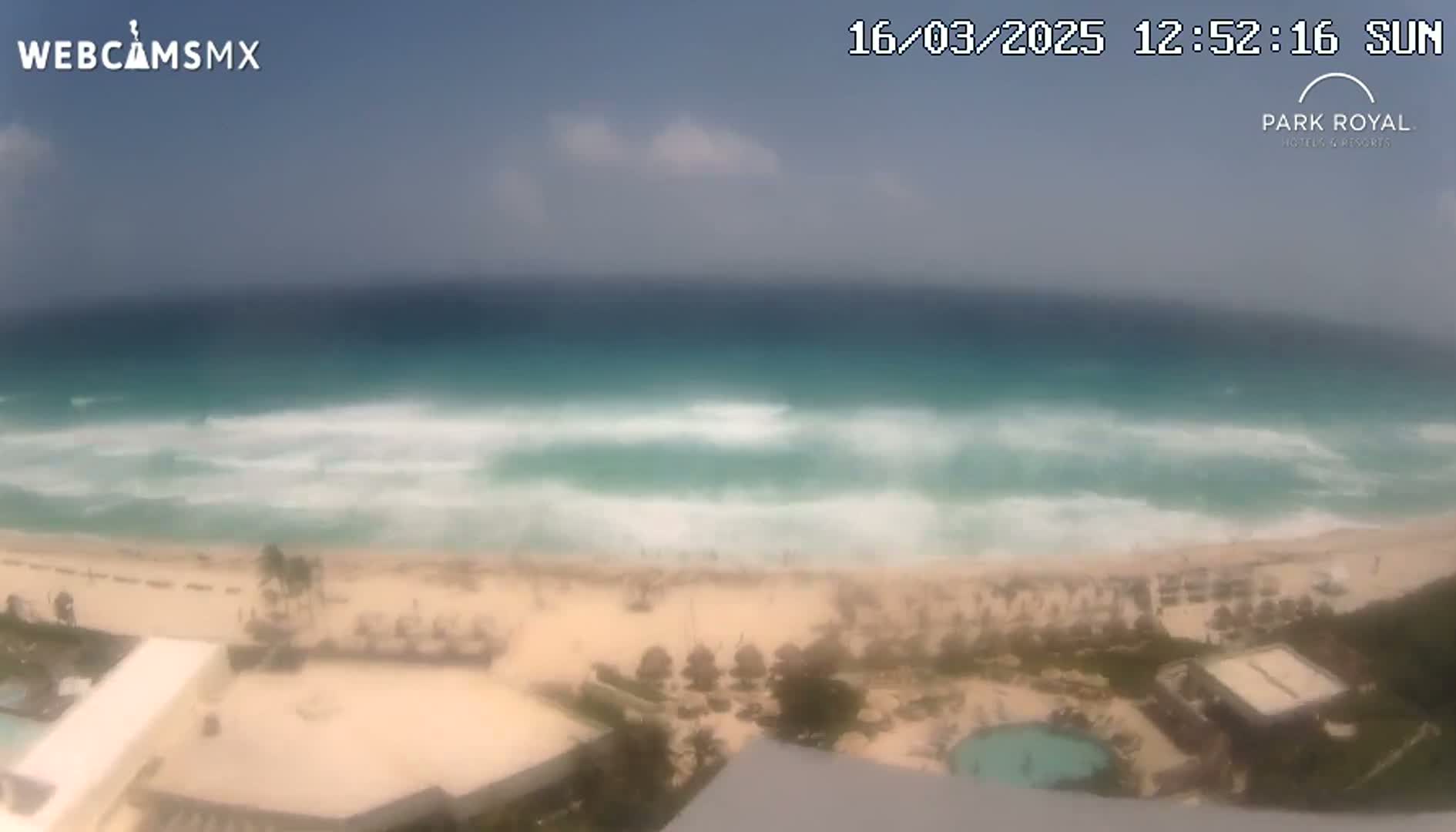 A mostly sunny day shows a view of turquoise ocean waves breaking on a white sand beach, with resort buildings and a swimming pool visible in the foreground.