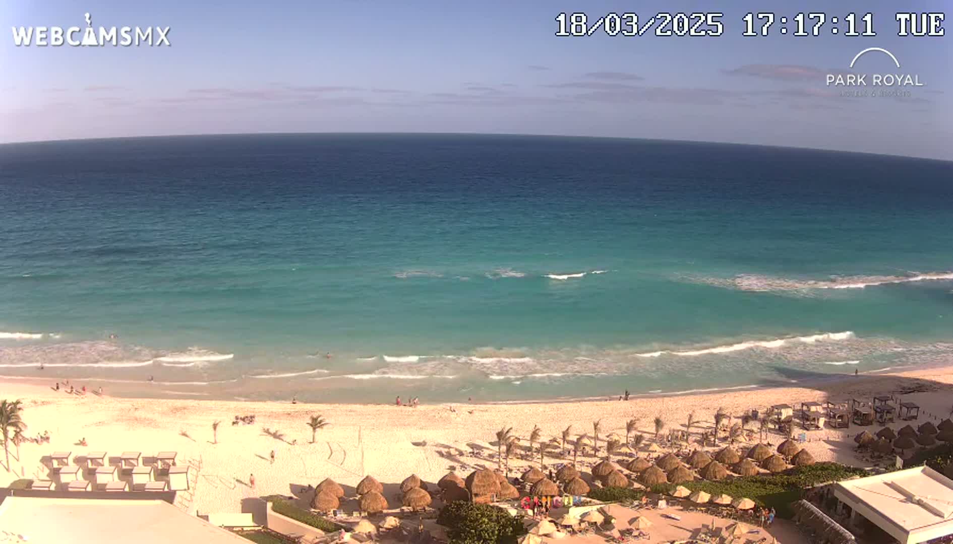 A beach with turquoise water and gentle waves under a clear sky, showing a resort with numerous thatched umbrellas and beach chairs.