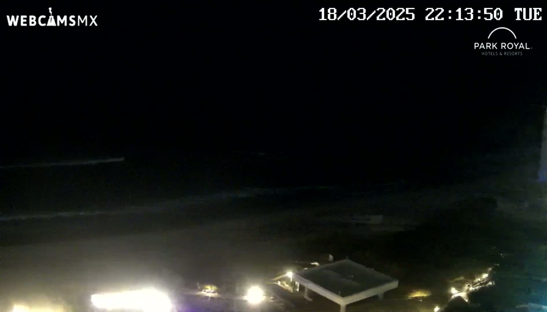 At night, a dark ocean with small waves is visible from a high vantage point, with lights illuminating a beach area and nearby structures.