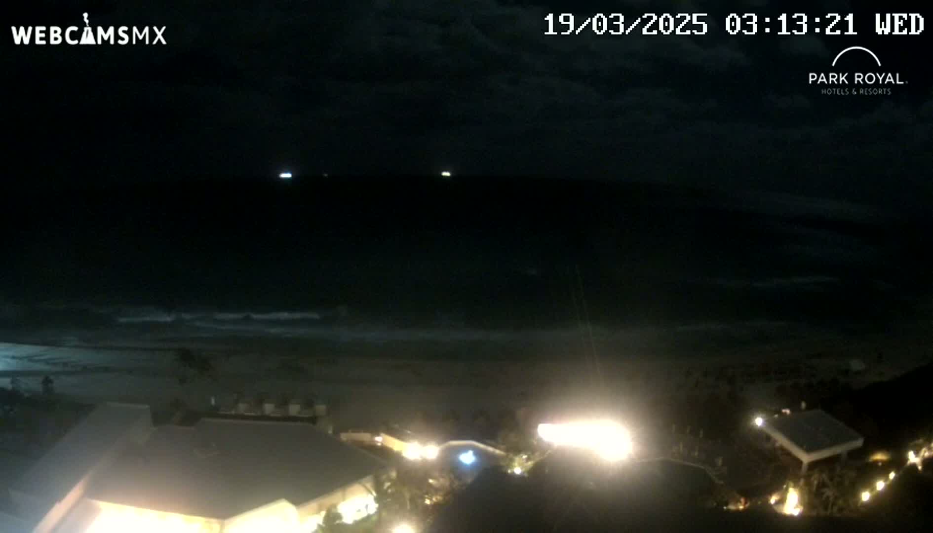 A nighttime view of a dark ocean with gentle waves lapping a beach, and illuminated buildings are visible in the foreground under a cloudy sky.