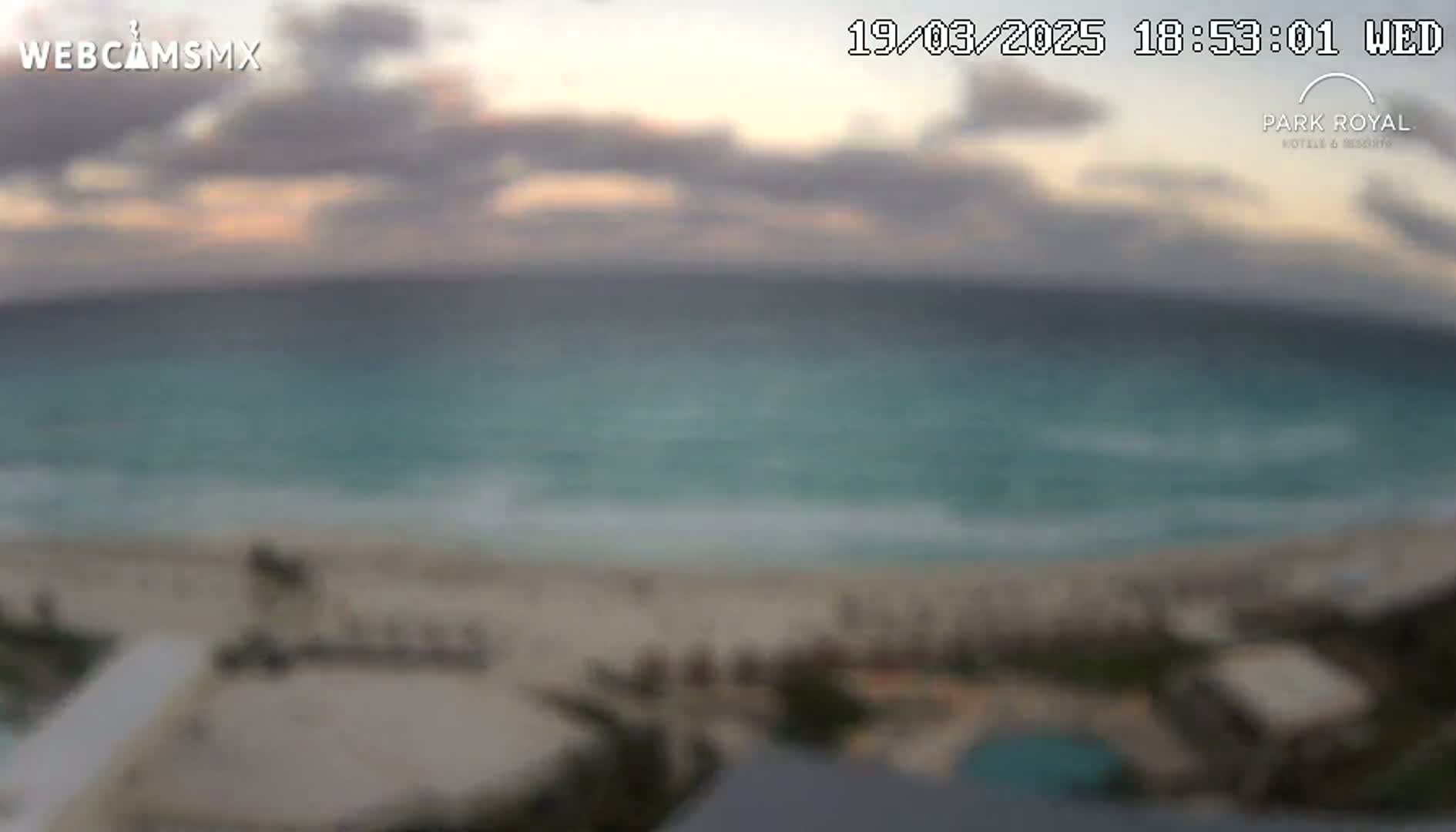 A blurry, high-angle view shows a beach with turquoise water under a partly cloudy sky at dusk.