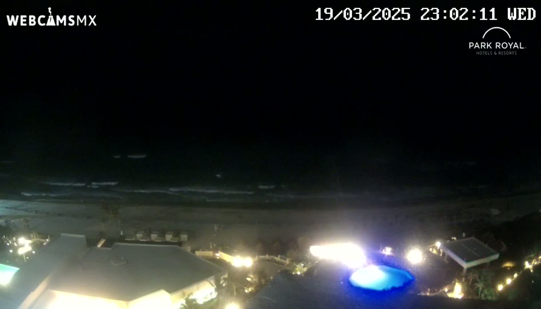 A nighttime view of a beach with gently rolling waves, seen from above a resort with illuminated buildings and a swimming pool.