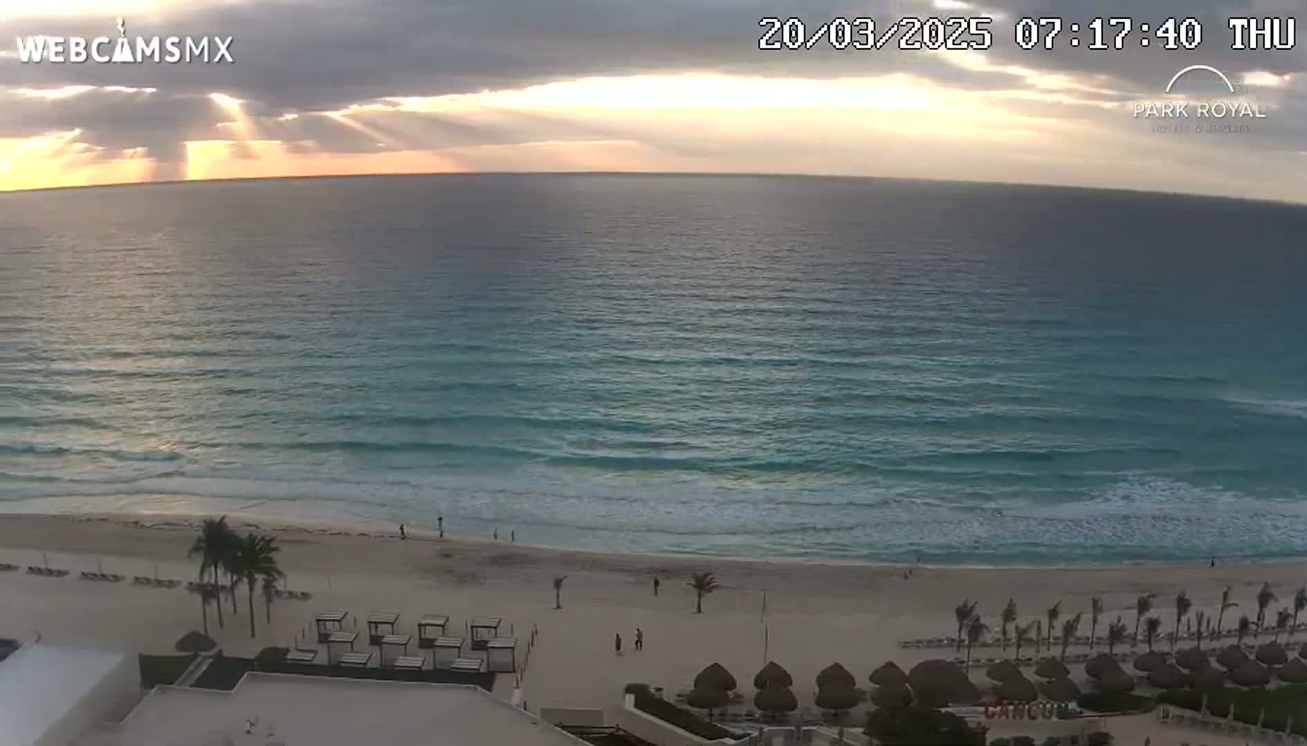 Sunbeams break through cloudy skies over a tranquil ocean, lapping gently at a sandy beach lined with palm trees and beach umbrellas.
