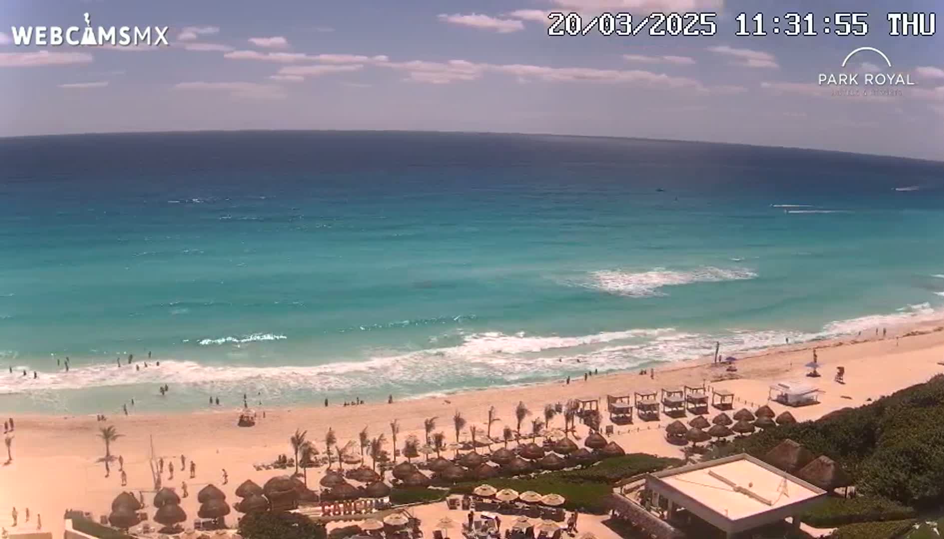 A partly cloudy day shows a beach with turquoise water, gentle waves, and people swimming and relaxing on the sand and under beach umbrellas.