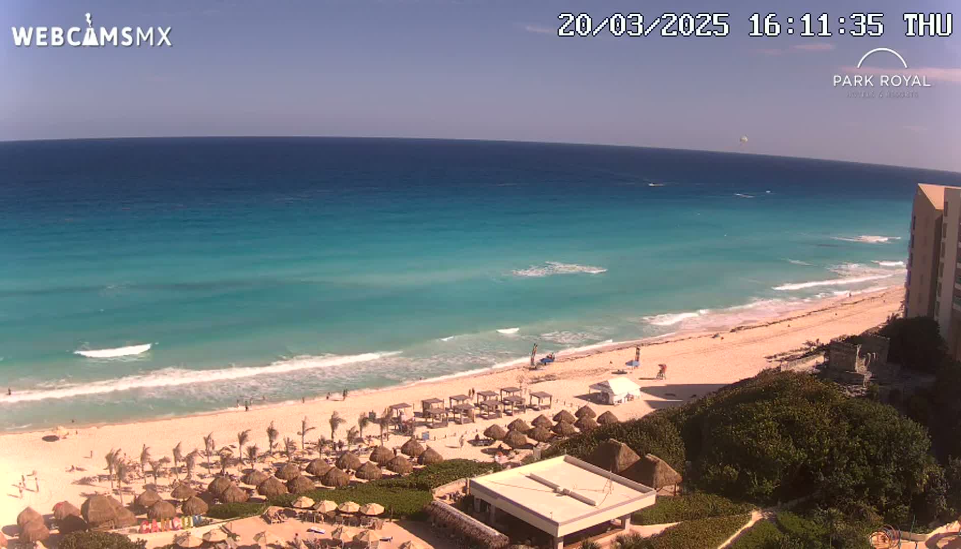 A beach with turquoise water and gentle waves under a clear blue sky, featuring numerous beach umbrellas and a resort building.