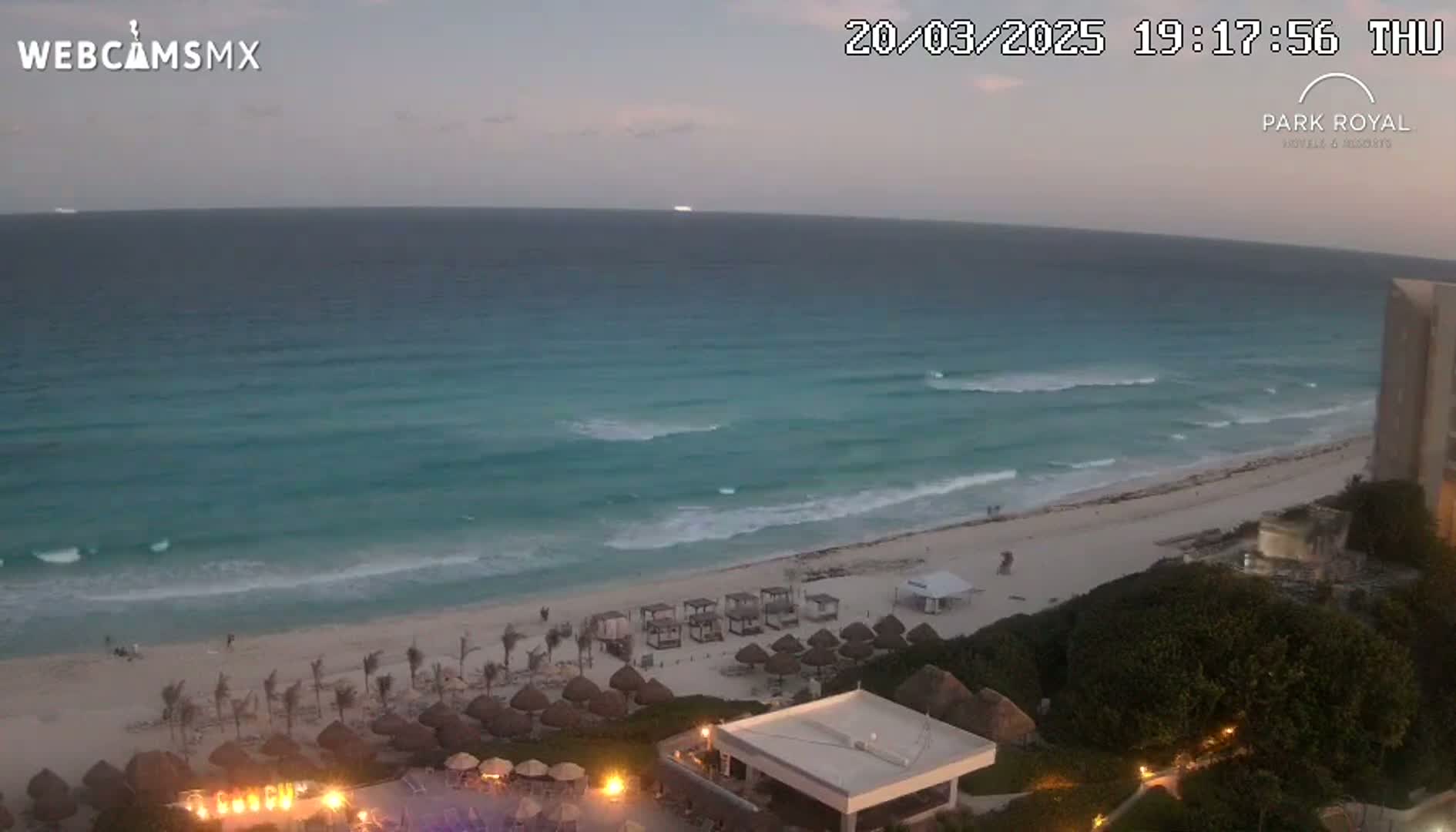 A beach at dusk with gentle waves lapping the shore, overlooked by a resort with lights illuminating its grounds.