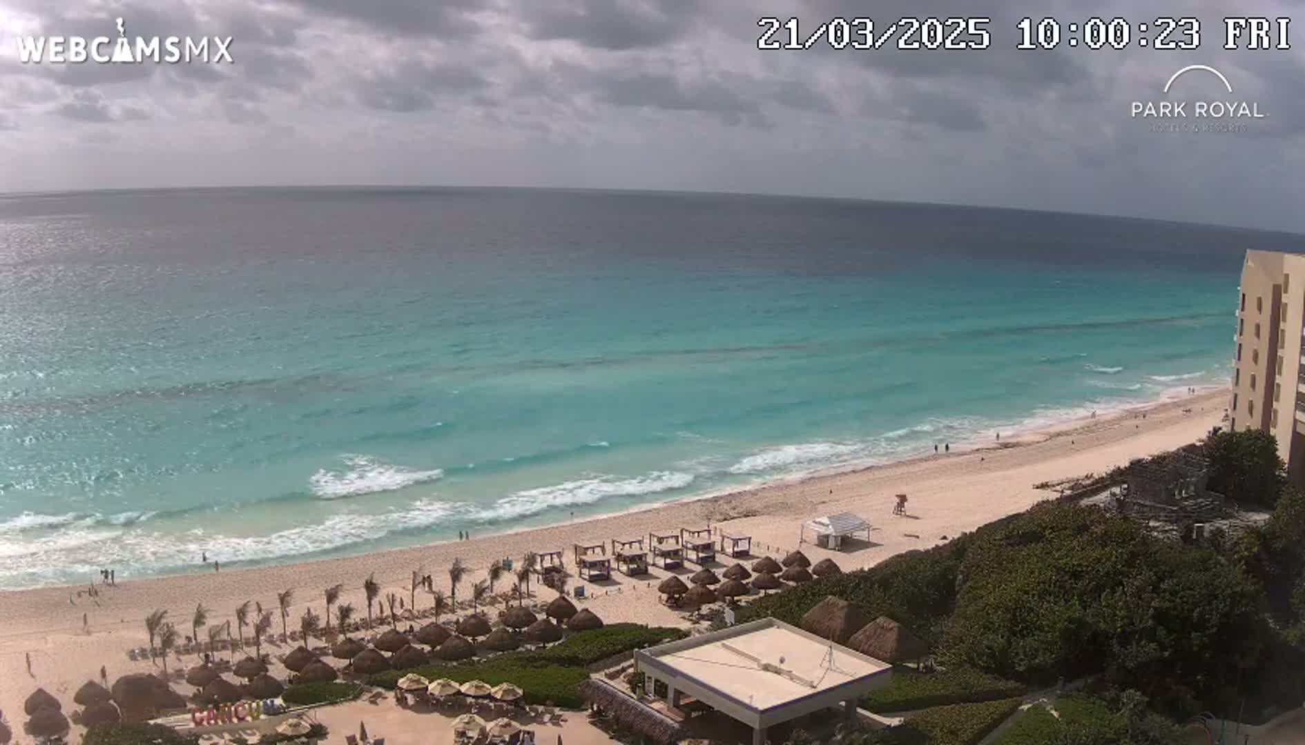 An expansive view of a sandy beach with turquoise water and small waves under a partly cloudy sky shows beach umbrellas and a resort building.
