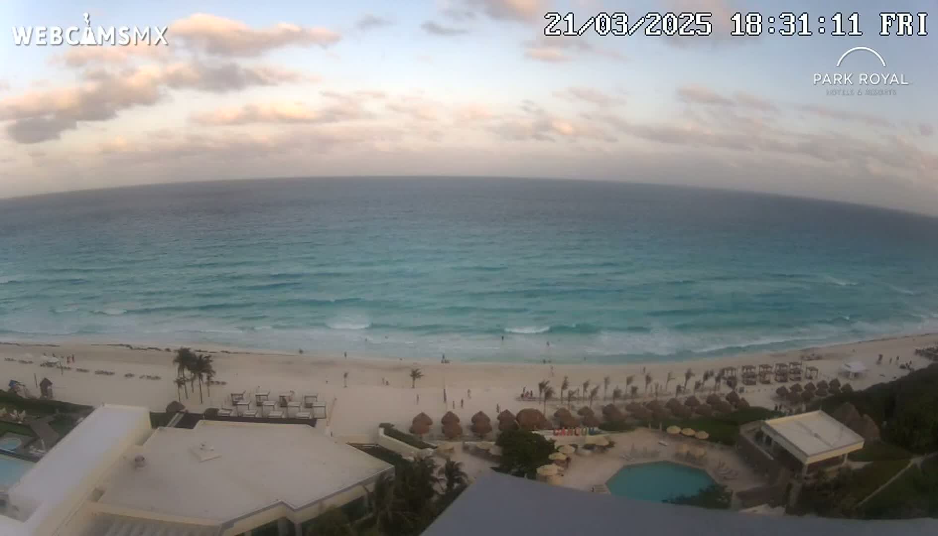 An ocean beach with light waves under a partly cloudy sunset sky is viewed from above a resort with a swimming pool.