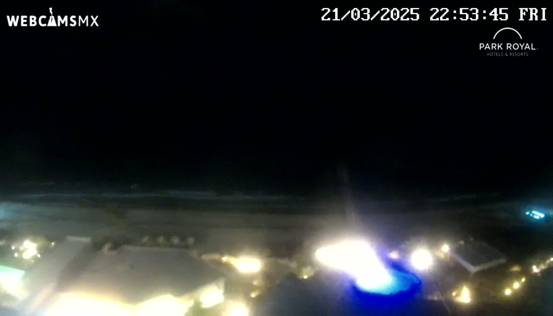 A nighttime view of a beach and resort area with a lit-up pool, showing calm ocean waves under a dark sky.