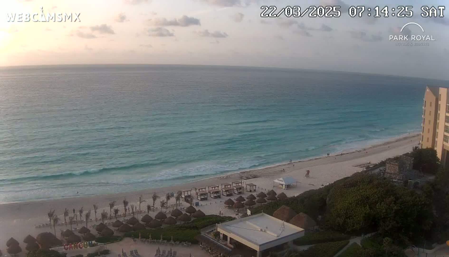 A mostly clear sunrise illuminates a beach with calm waves and rows of thatched-roof structures.