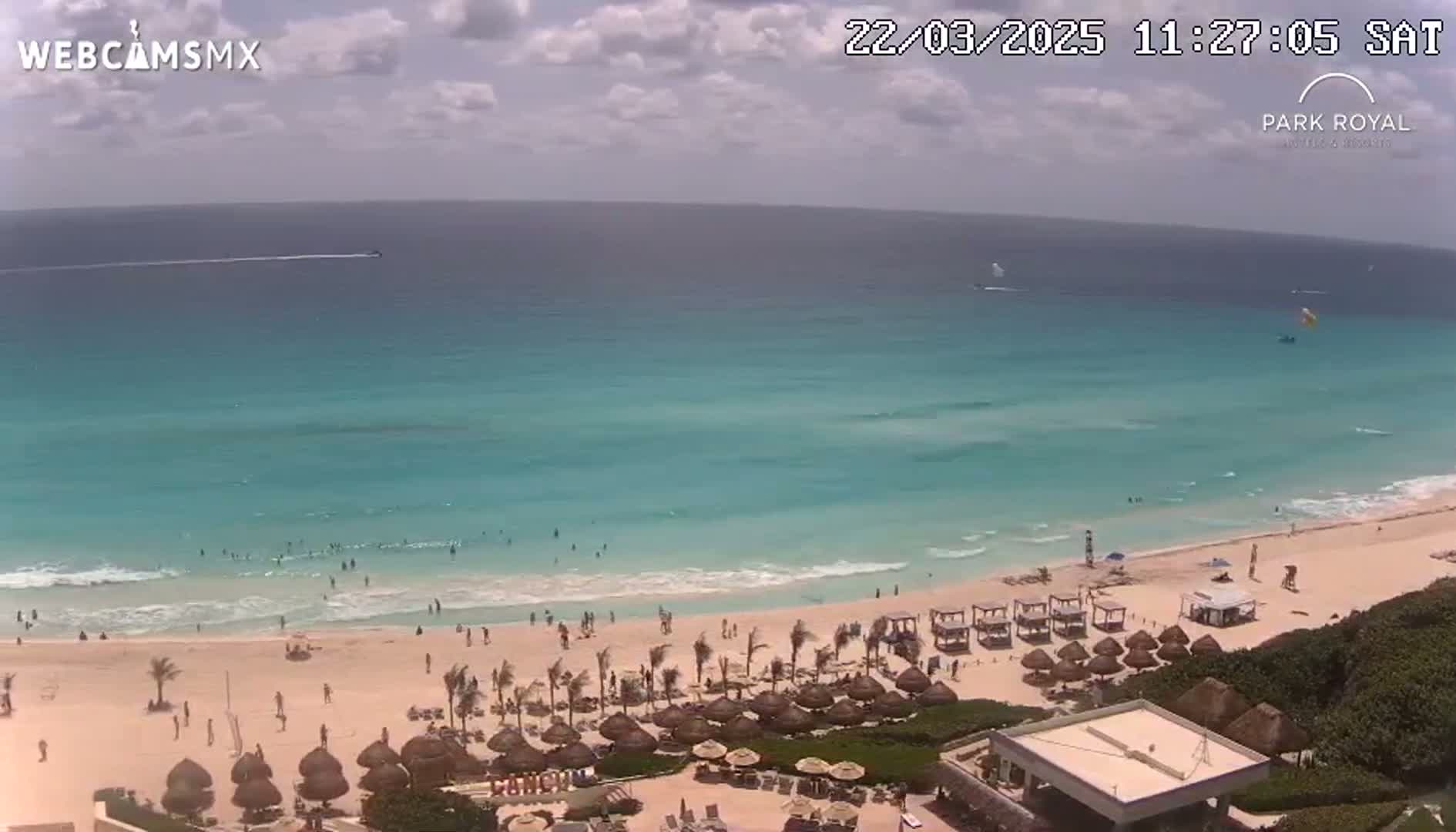 A beach scene shows turquoise water lapping a sandy shore where people are swimming and relaxing under umbrellas, with partly cloudy skies overhead.