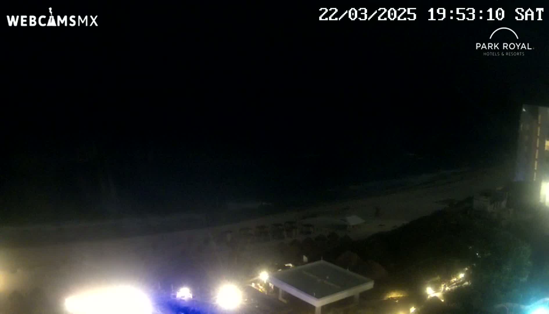 A nighttime beach scene shows dark water lapping a sandy shore, with lights illuminating a section of beach and nearby buildings.