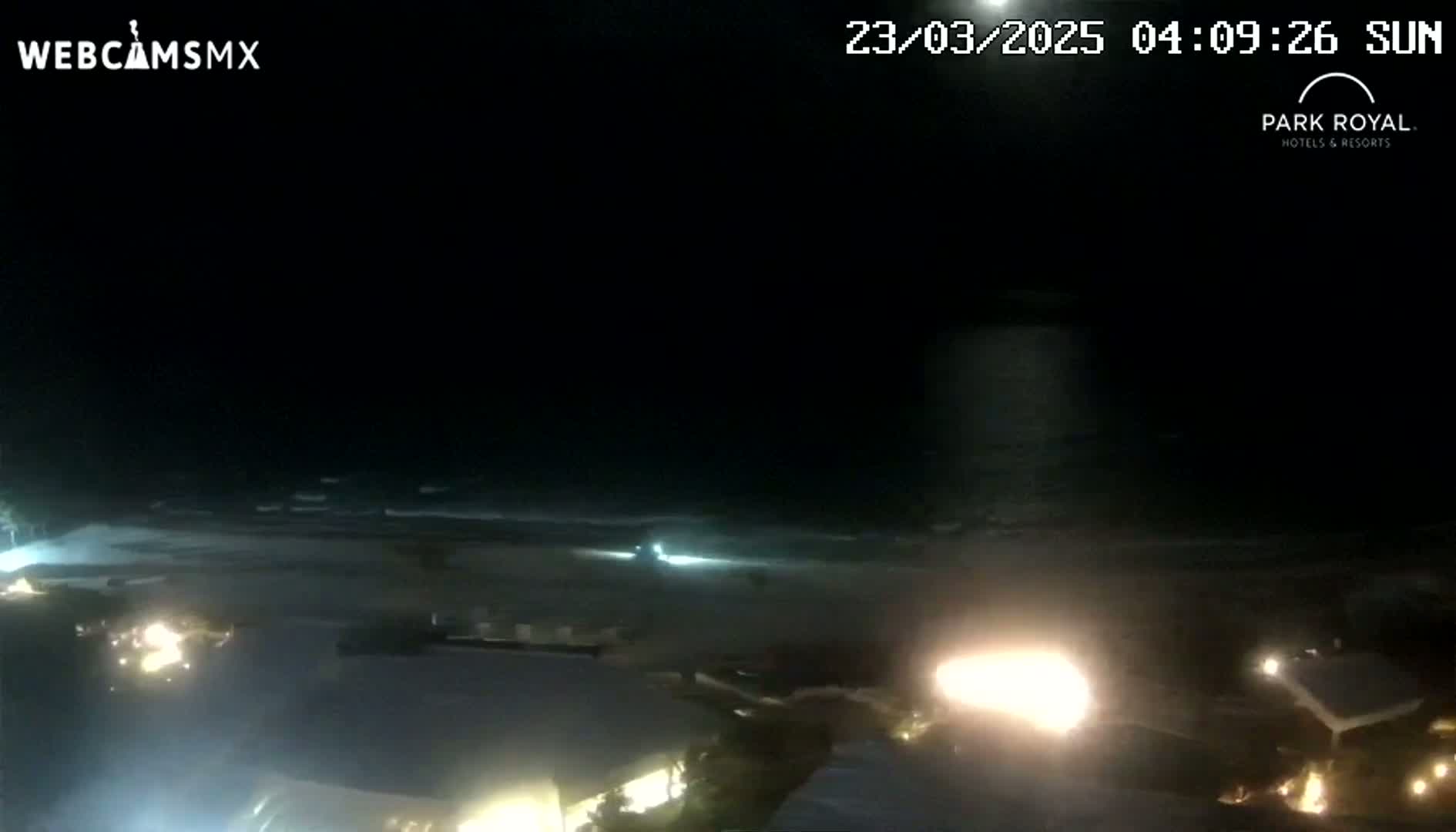 A nighttime beach scene shows dark, gently waving water under a clear, moonlit sky, with illuminated buildings and vehicles in the foreground.