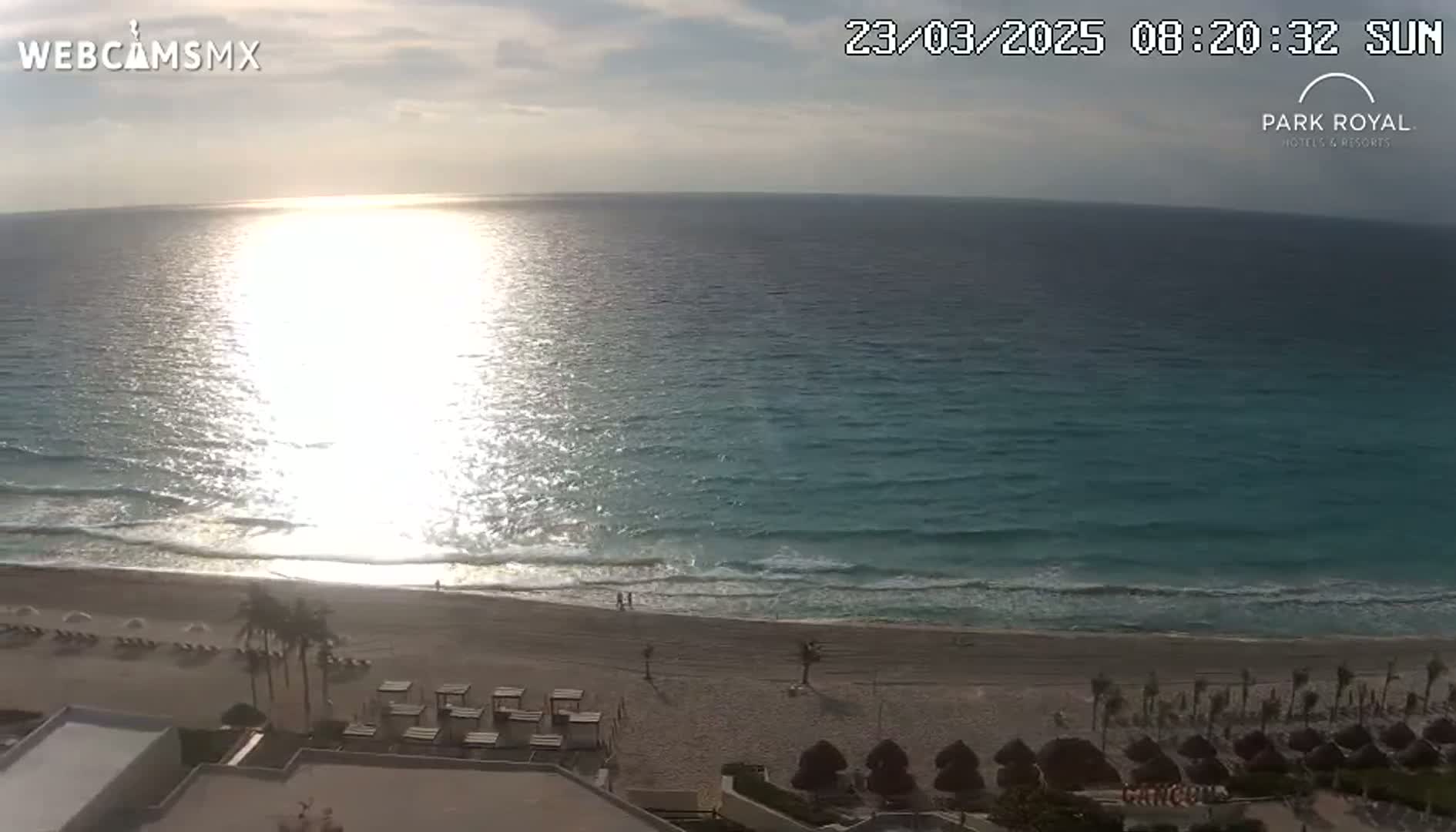A mostly sunny day shows a beach with calm waves and a bright, reflective ocean.