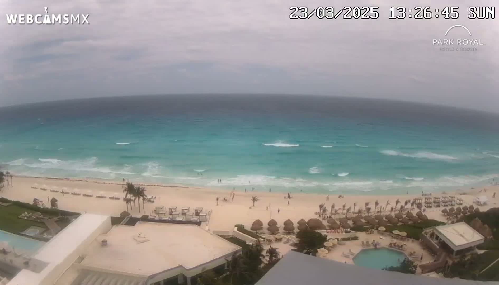 An overhead view shows a beach with turquoise water and small waves under an overcast sky, with resort buildings and a swimming pool visible in the foreground.