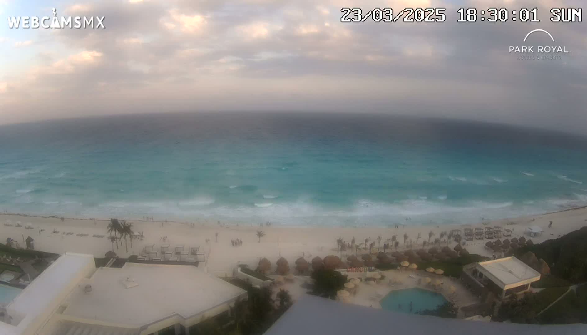 An aerial view shows a beach with turquoise water and small waves under a partly cloudy sunset sky.