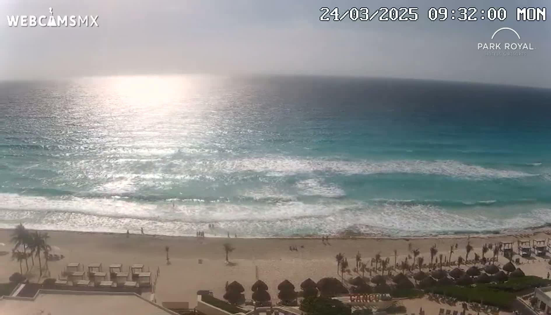 A mostly sunny day at a beach with turquoise water and small waves breaking on a sandy shore, showing a beach resort in the foreground.