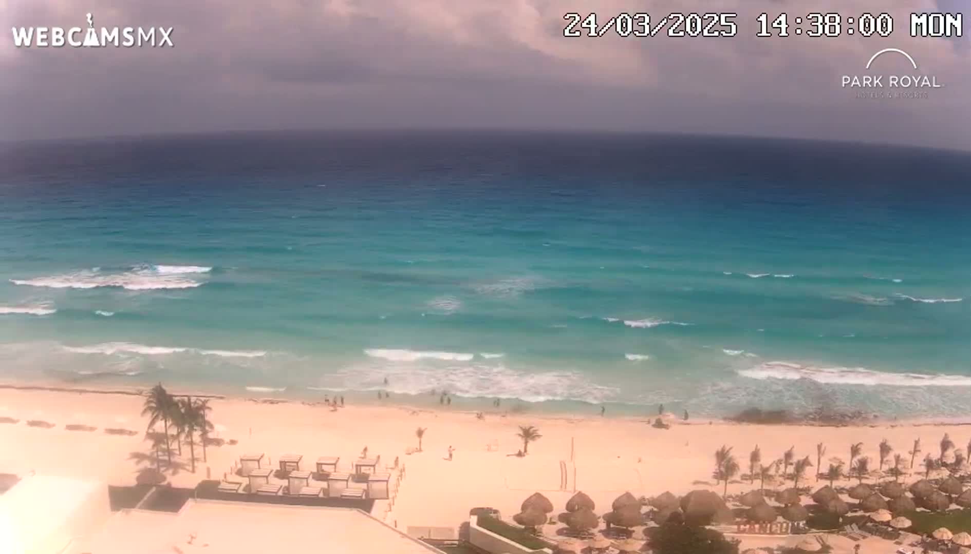 An elevated view shows a partly cloudy sky over a beach with turquoise water and gentle waves, and some people are visible on the shore near resort-style umbrellas and cabanas.