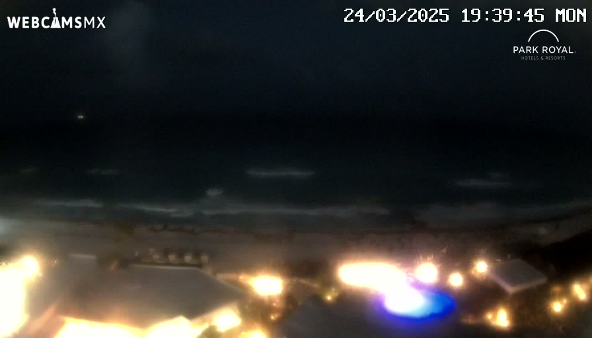 A nighttime view of a dark ocean with gently rolling waves, a sandy shore, and illuminated buildings and a pool in the foreground.