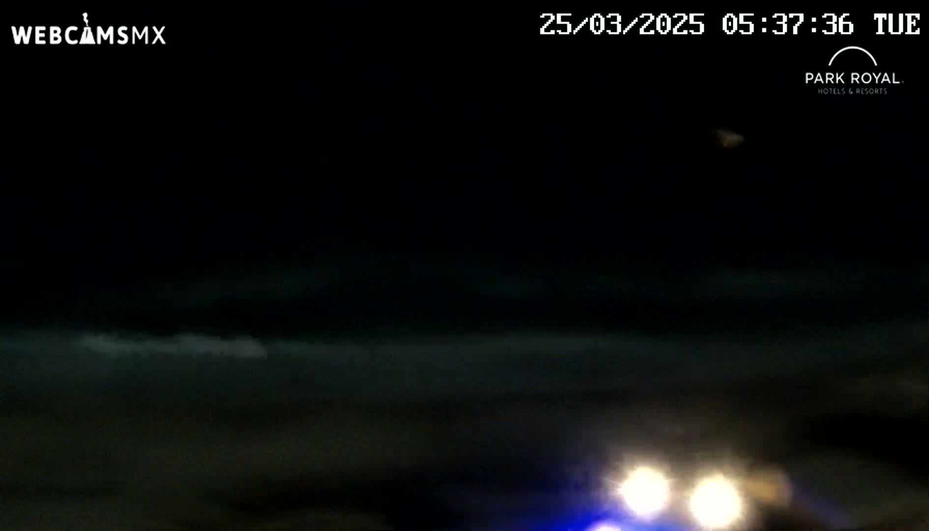 At night, a dark body of water is visible under a mostly clear, dark sky, with the headlights of a vehicle approaching in the bottom right corner.