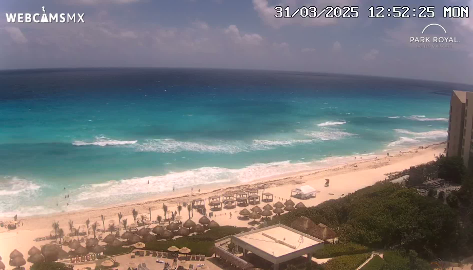 A partly cloudy day shows turquoise water with waves breaking on a sandy beach lined with thatched umbrellas and a building.
