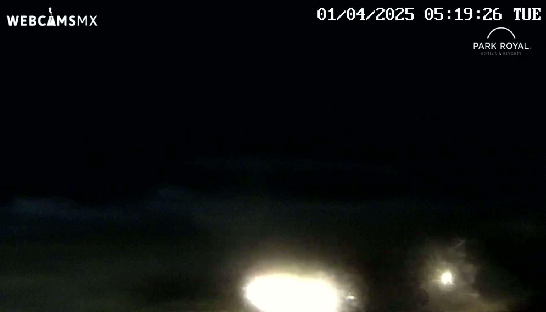 A nighttime, dark scene with a bright light source in the lower center and fainter lights in the lower right corner, under a cloudy sky.