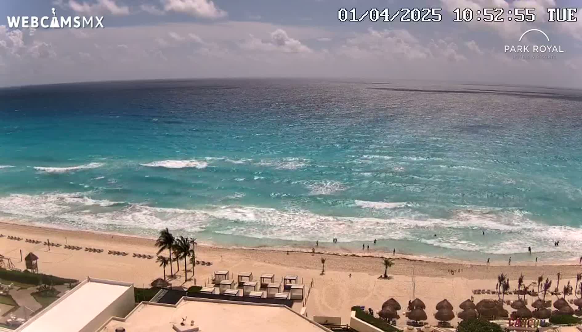 A mostly sunny day shows turquoise water with moderate waves lapping a sandy beach with beachgoers and resort amenities.