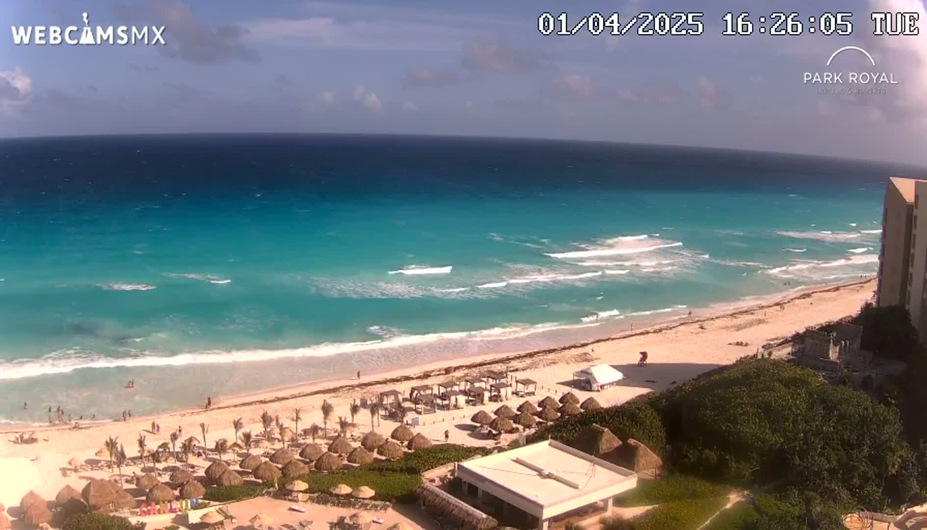 A beach with turquoise water and small waves under a mostly sunny sky is lined with palm trees and beach umbrellas, next to a resort building.