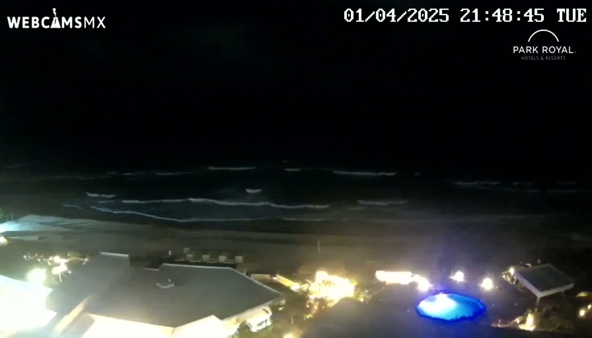 A nighttime view of a beach with gently rolling waves, illuminated buildings, and a pool with blue lighting.