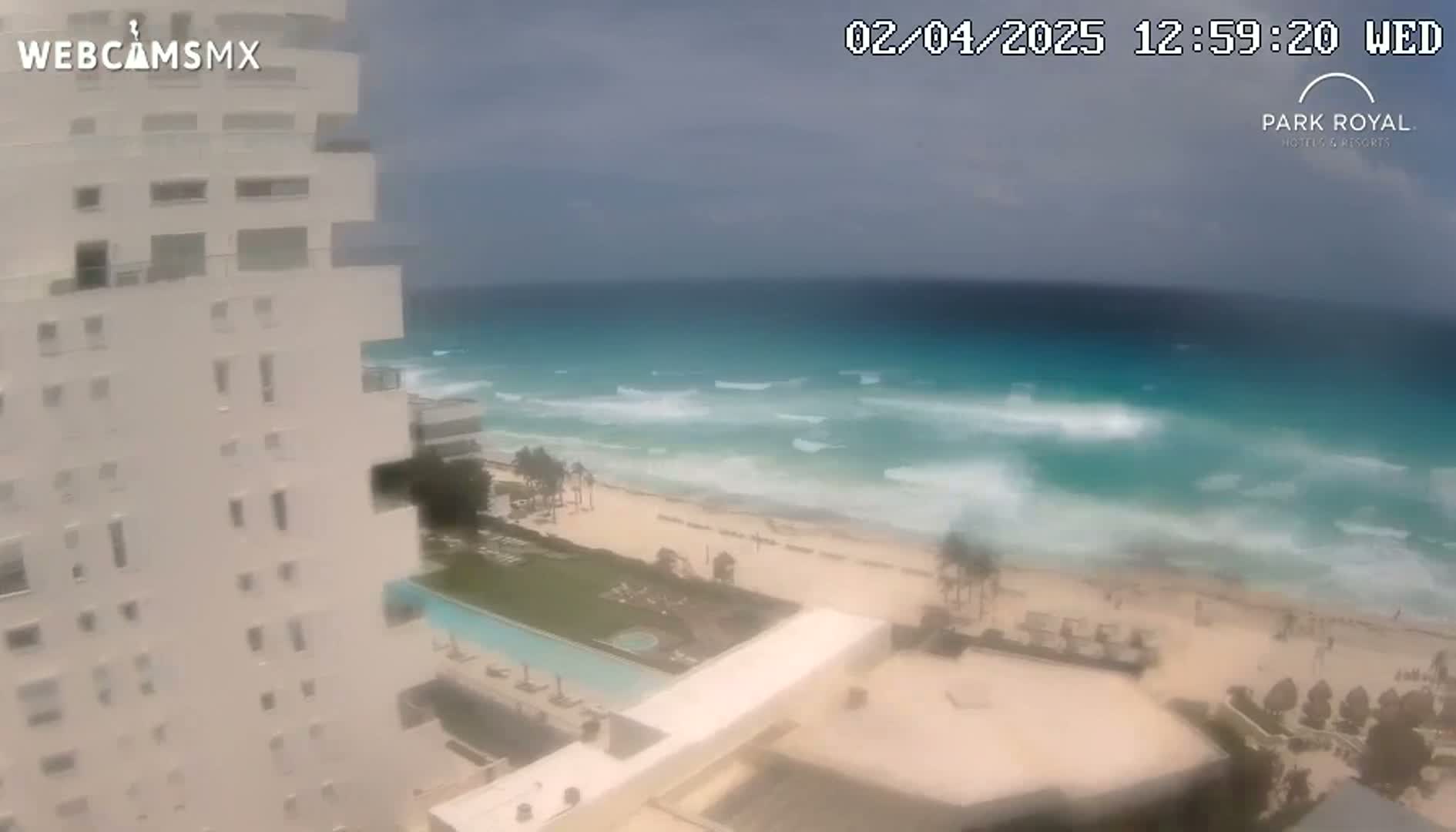 A high-angle view shows a white building overlooking a beach with turquoise water and moderate waves under an overcast sky.
