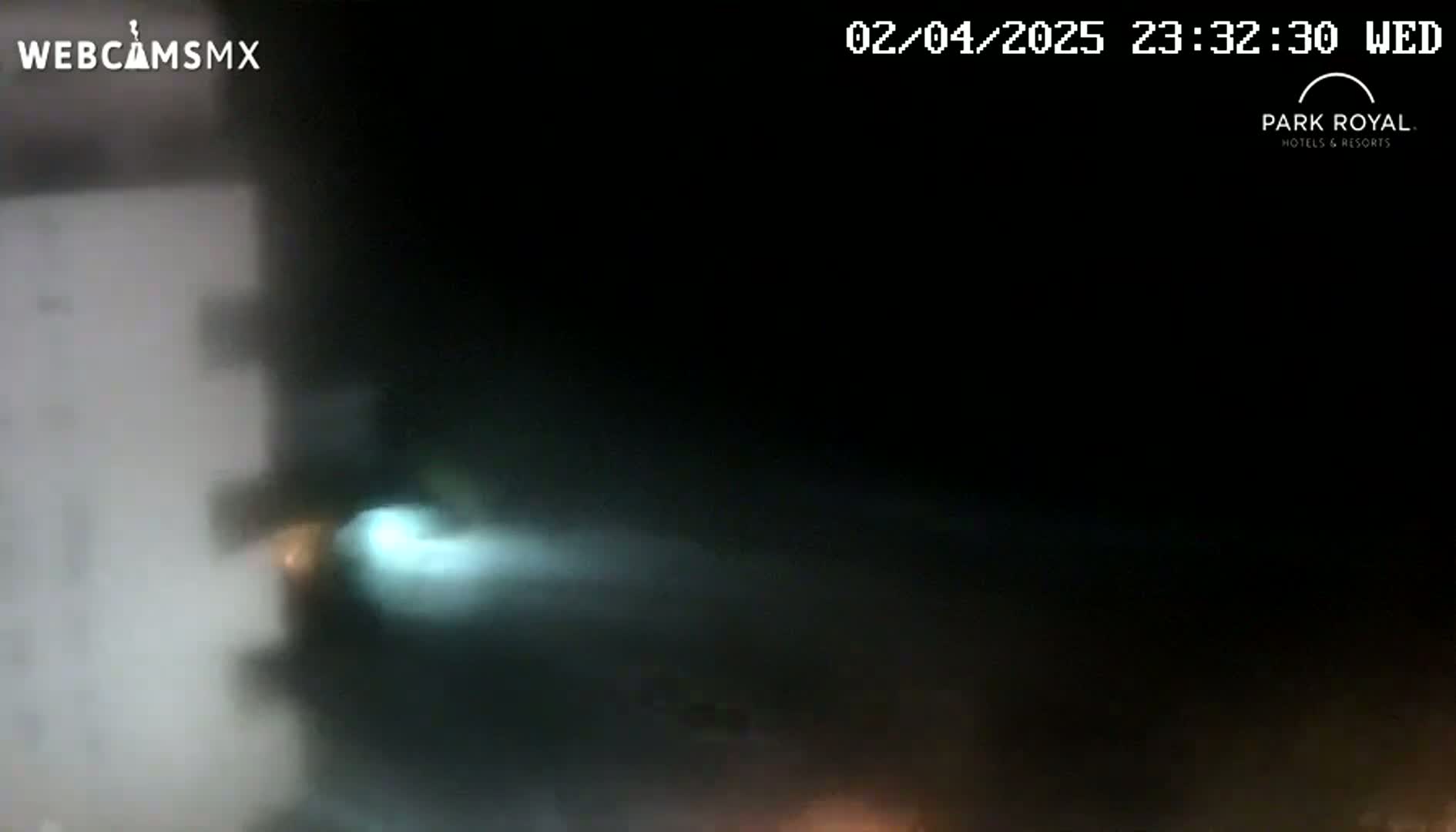 A blurry nighttime image shows a bright light streaking across a dark sky, partially obscuring a light-colored building on the left.