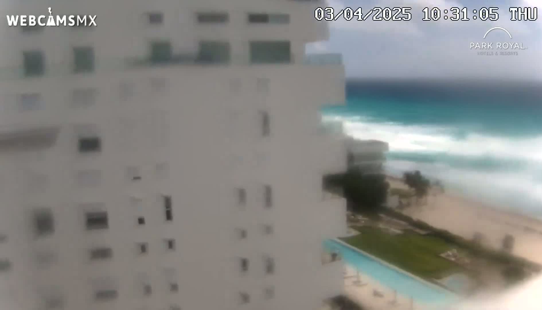 A white building overlooks a beach with turquoise water and moderately large waves under partly cloudy skies.
