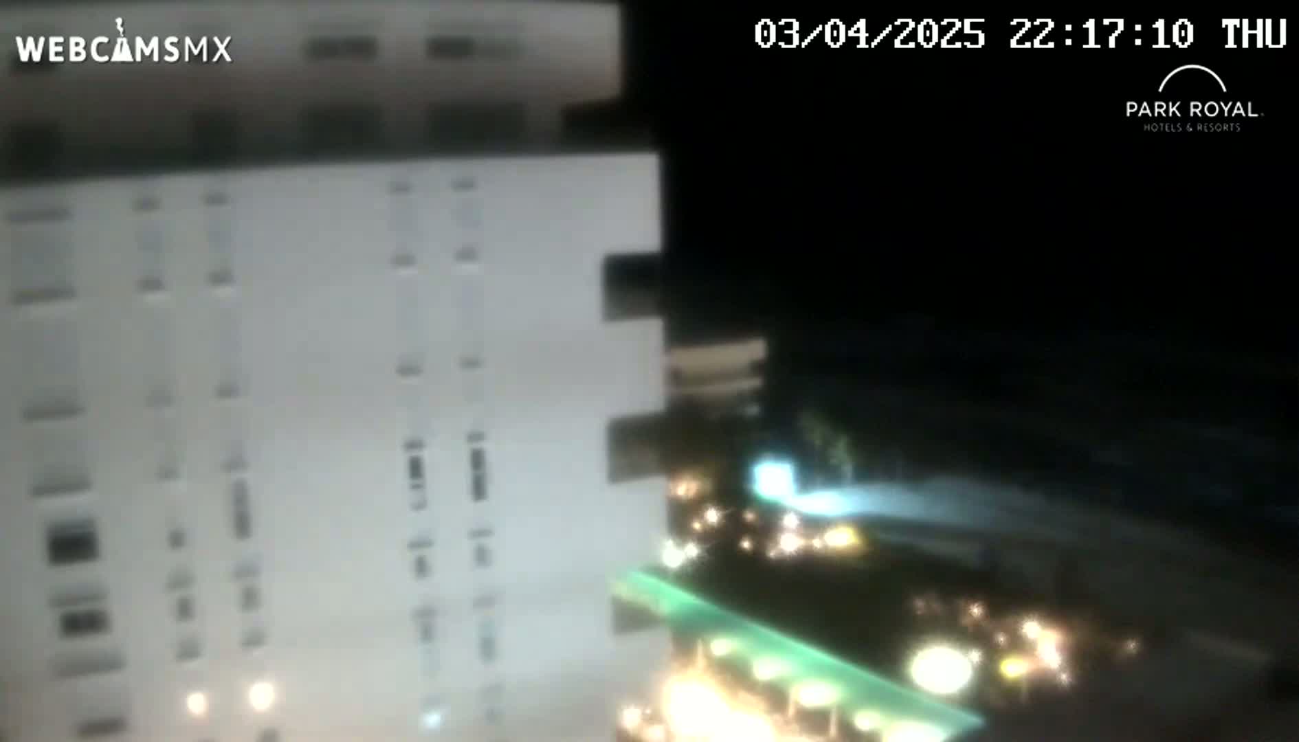 At night, a brightly lit, multi-story building is partially visible, with other illuminated buildings and vegetation in the foreground.
