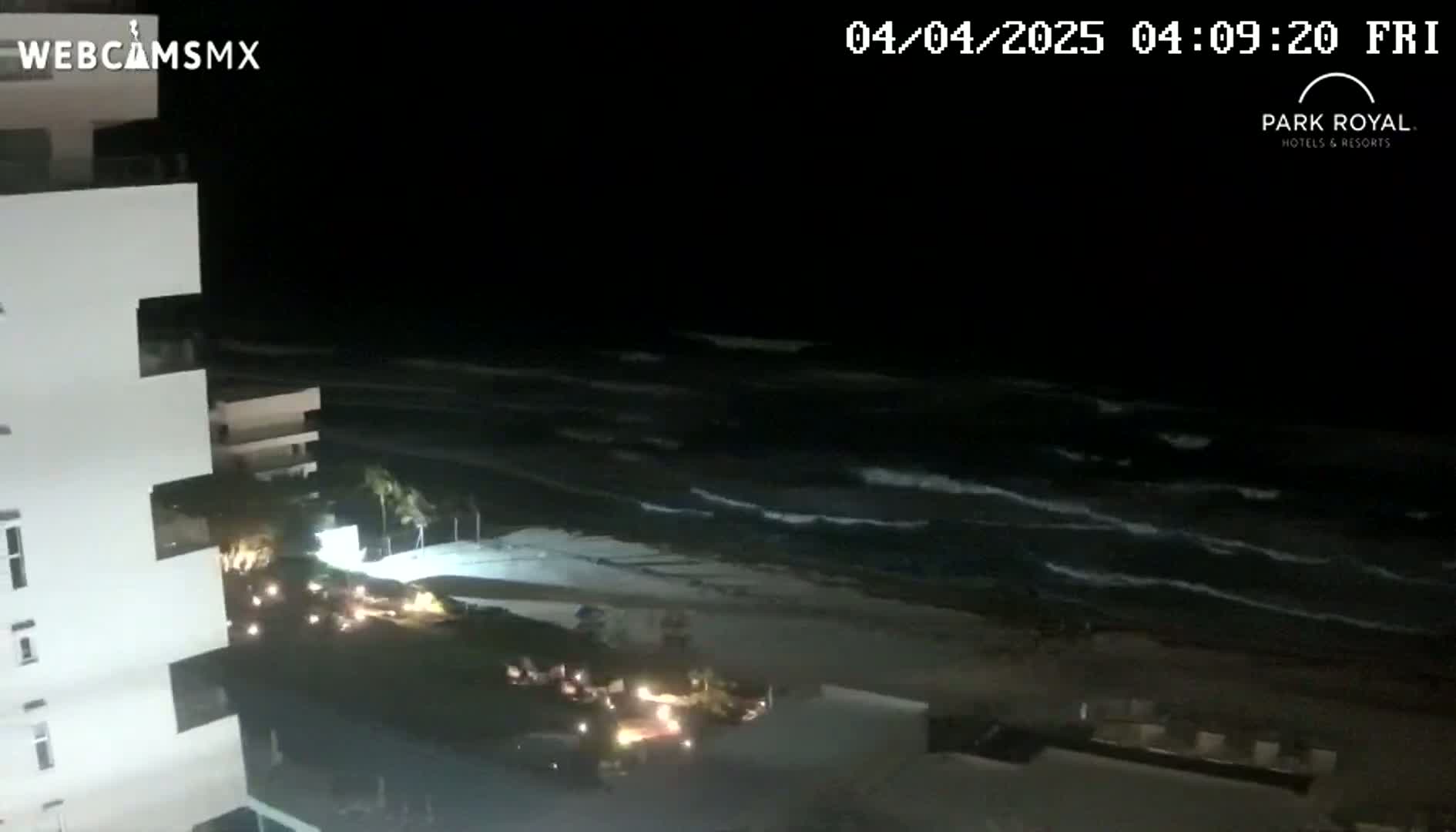 A nighttime view shows a beach with moderate waves under a dark sky, next to a tall building with lights visible near the ground floor.