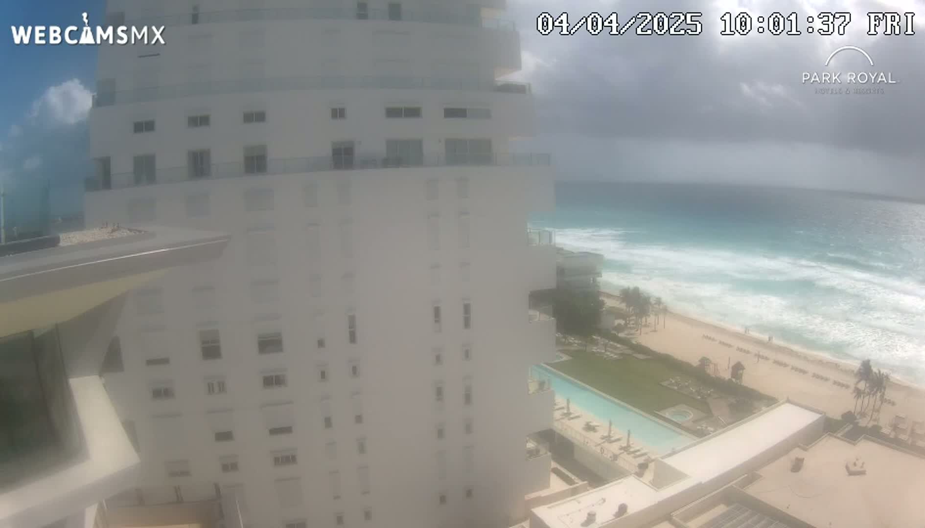 A hazy, partly cloudy day shows a white high-rise building overlooking a beach with turquoise water and breaking waves.