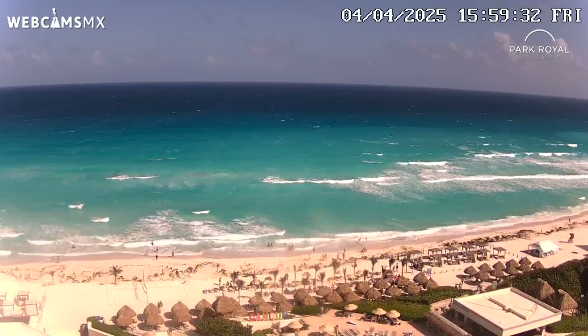 A sandy beach with turquoise water and small waves under a mostly clear sky, featuring beach umbrellas and resort buildings.