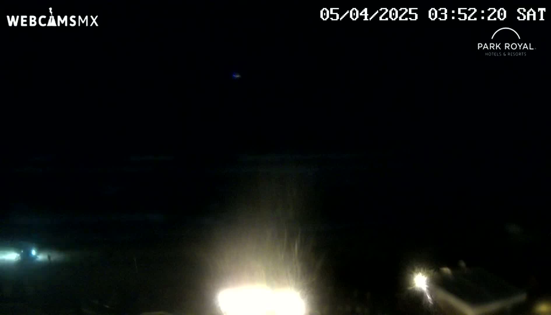 A nighttime, dark scene shows a bright light in the lower center, with faint lights visible in the distance, and a dark body of water in the background under a clear sky.