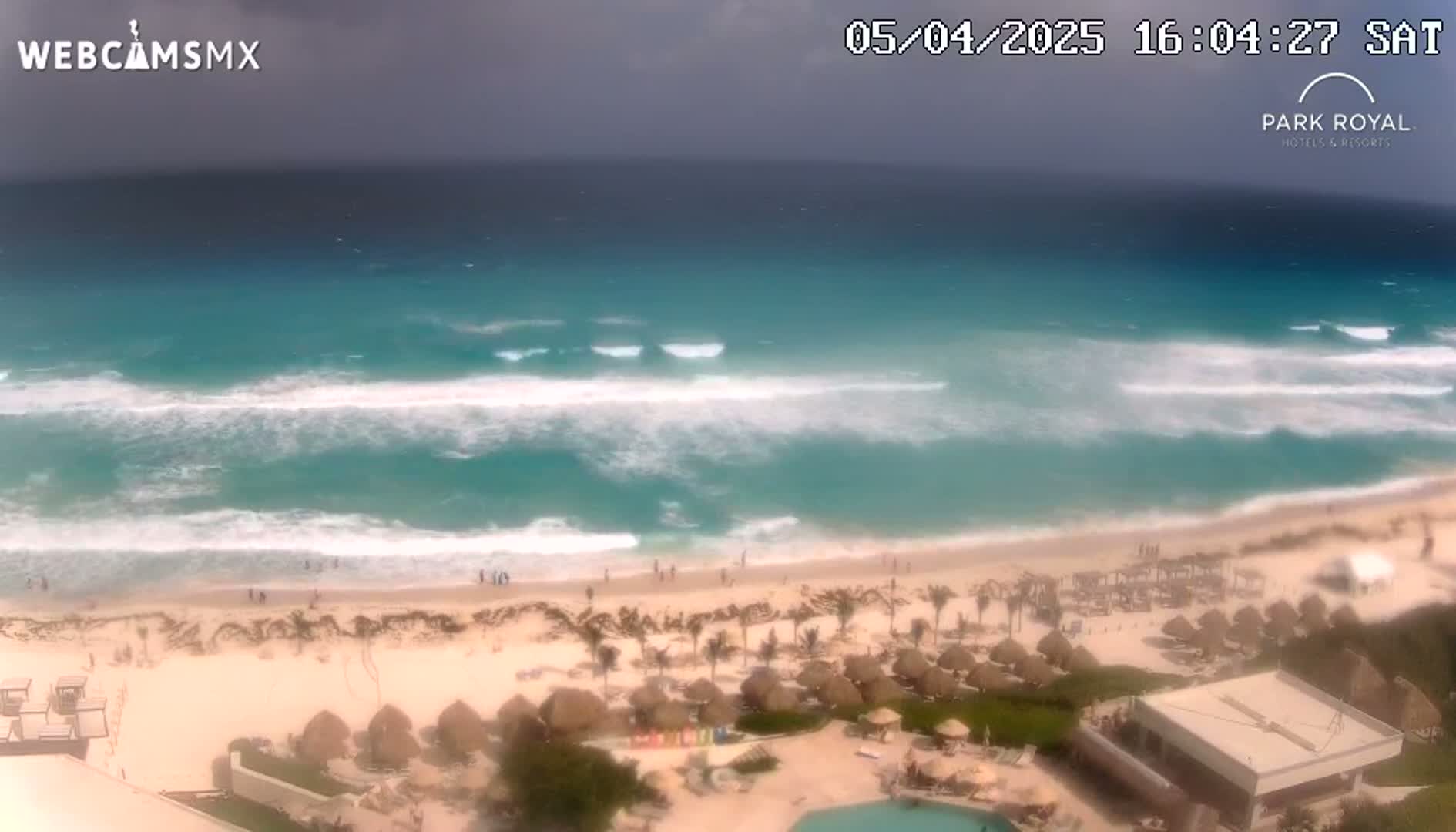 An elevated view shows a beach with turquoise water and moderate waves under an overcast sky;  palm trees and beach umbrellas are visible on the sand.