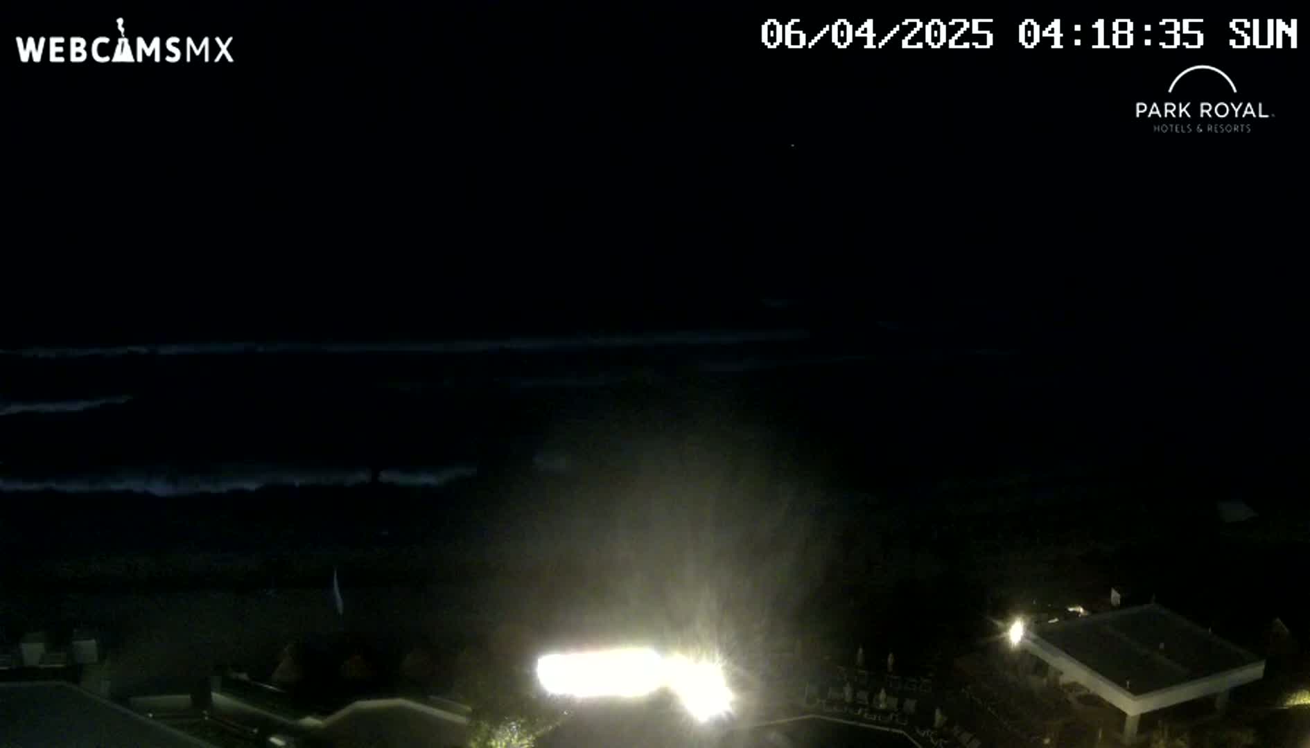 A nighttime, dark ocean with waves is visible in the distance, while brightly lit structures and landscaping are illuminated in the foreground.