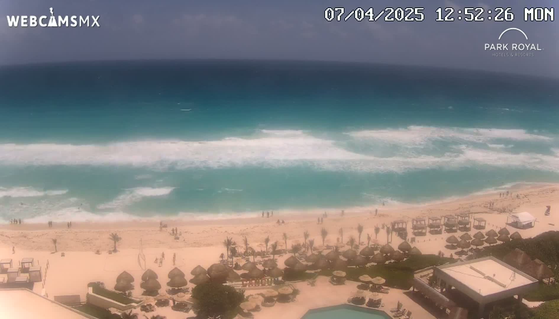 An aerial view shows a beach with turquoise water and moderate waves under a mostly sunny sky, with beach umbrellas and a resort visible.