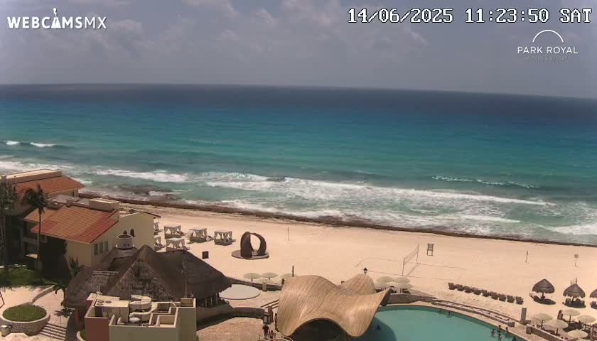 A beach resort with turquoise water, small waves, and a partly cloudy sky is shown from an elevated viewpoint.