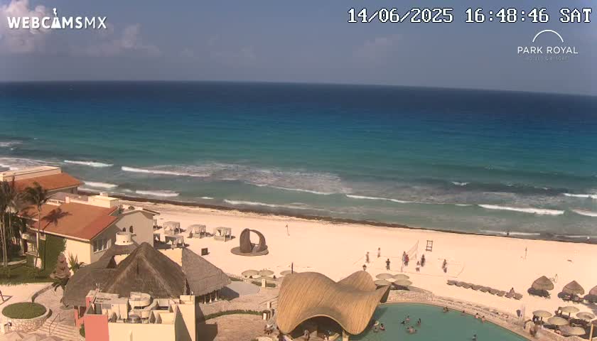 A beach resort with turquoise water, small waves, and a partly cloudy sky is shown from an elevated viewpoint.