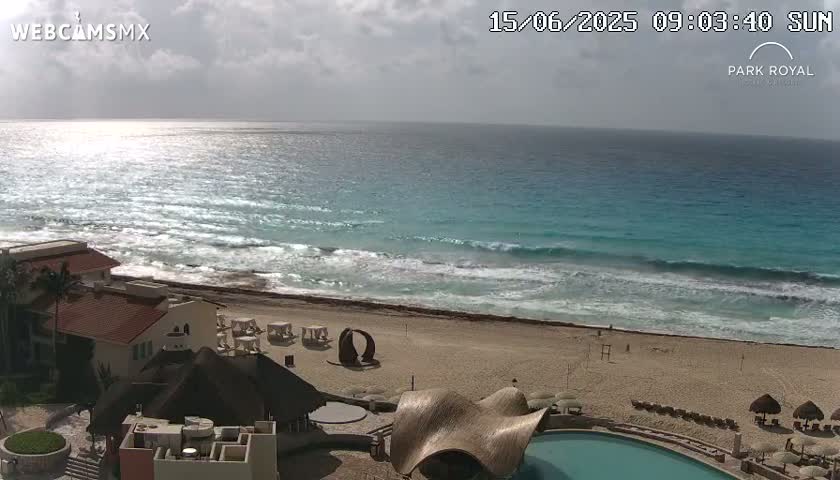 A partly sunny day shows turquoise ocean waves rolling onto a sandy beach with resort buildings and a swimming pool.