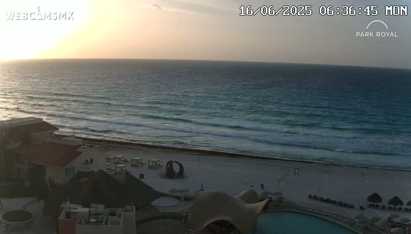 A sunrise over a calm ocean with small waves breaking on a sandy beach, seen from a high vantage point overlooking beachside buildings.