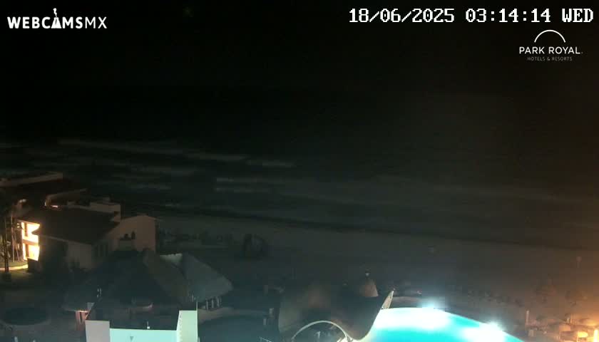 A nighttime view of a dark ocean with waves rolling onto a beach near buildings with lights on.