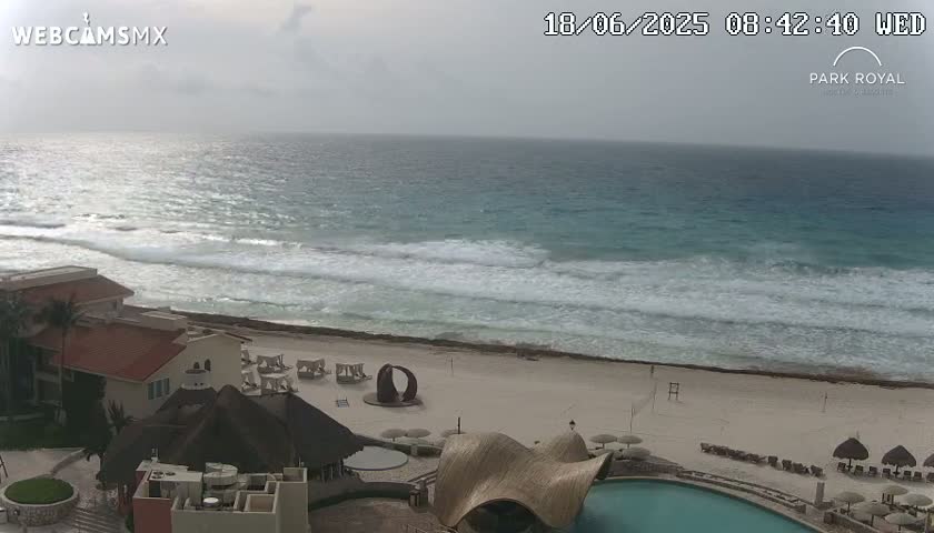 A mostly cloudy day shows a beach with moderate waves and a resort with a pool area.